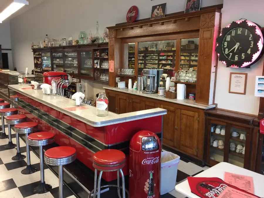 An old soda fountain with a white topped counter, red stools, black and white tiled floors and a wood hutch behind the counter.