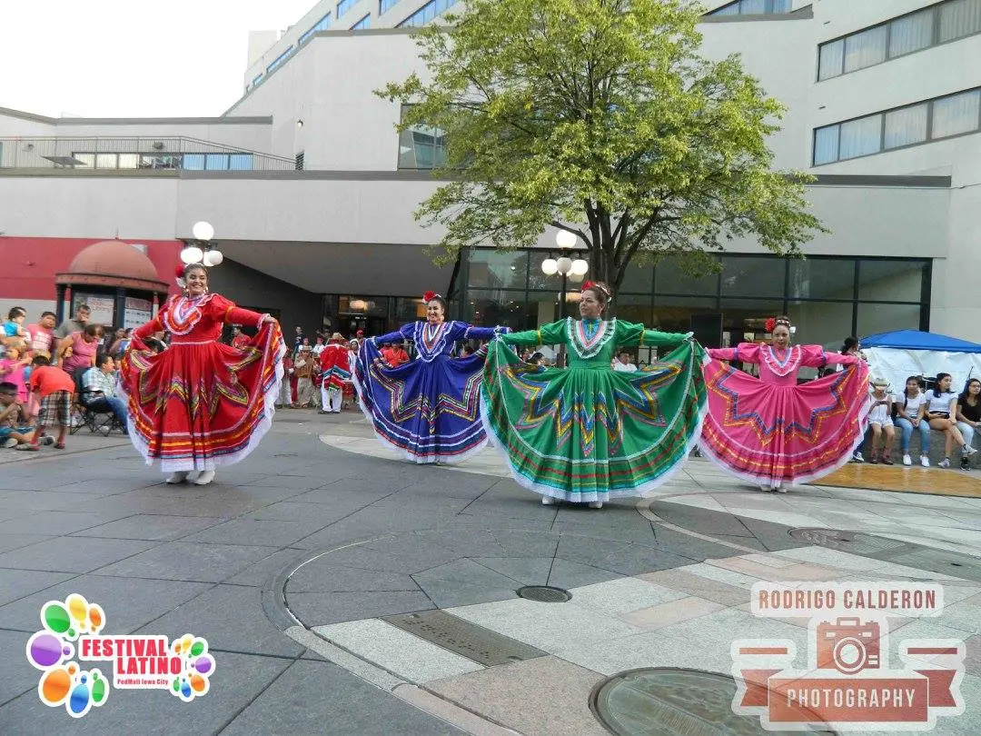Four Latina dances hold the skirts of their vibrantly colored dresses at shoulder height before a crowd.