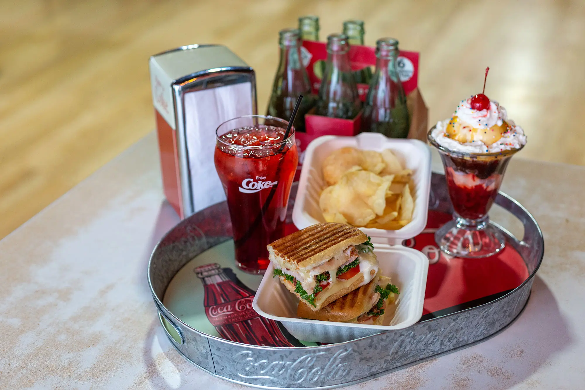 A tin tray containing a styrofoam box with a sandwich and chips alongside a coke and ice cream sundae.